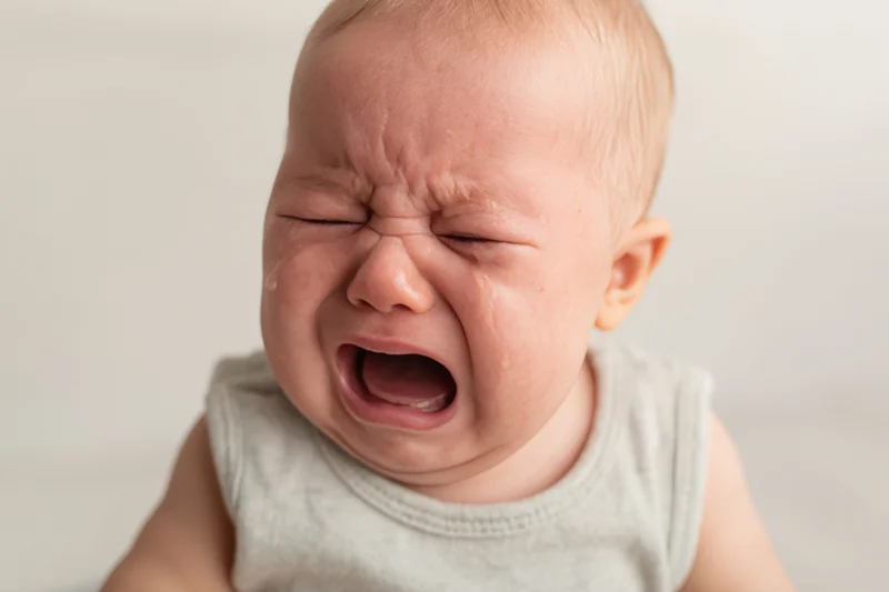 Crying baby with eyes squeezed shut and mouth open in a wail wearing a gray sleeveless top indoors