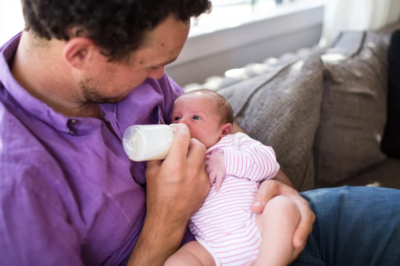 Father feeding newborn baby with bottle while sitting on couch at home