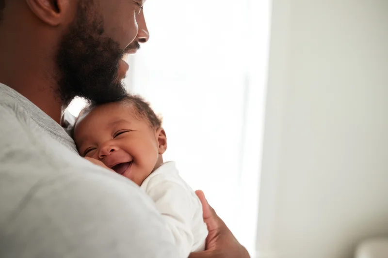 Father cradling newborn baby who is laughing while resting on his shoulder indoors