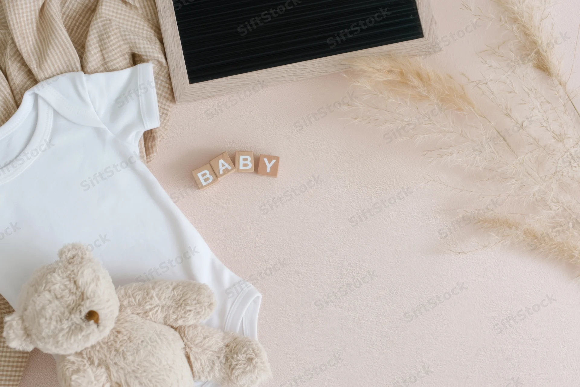 Baby items arranged in flat lay on pale wooden surface with onesie, teddy bear, letter blocks spelling baby, and pampas grass