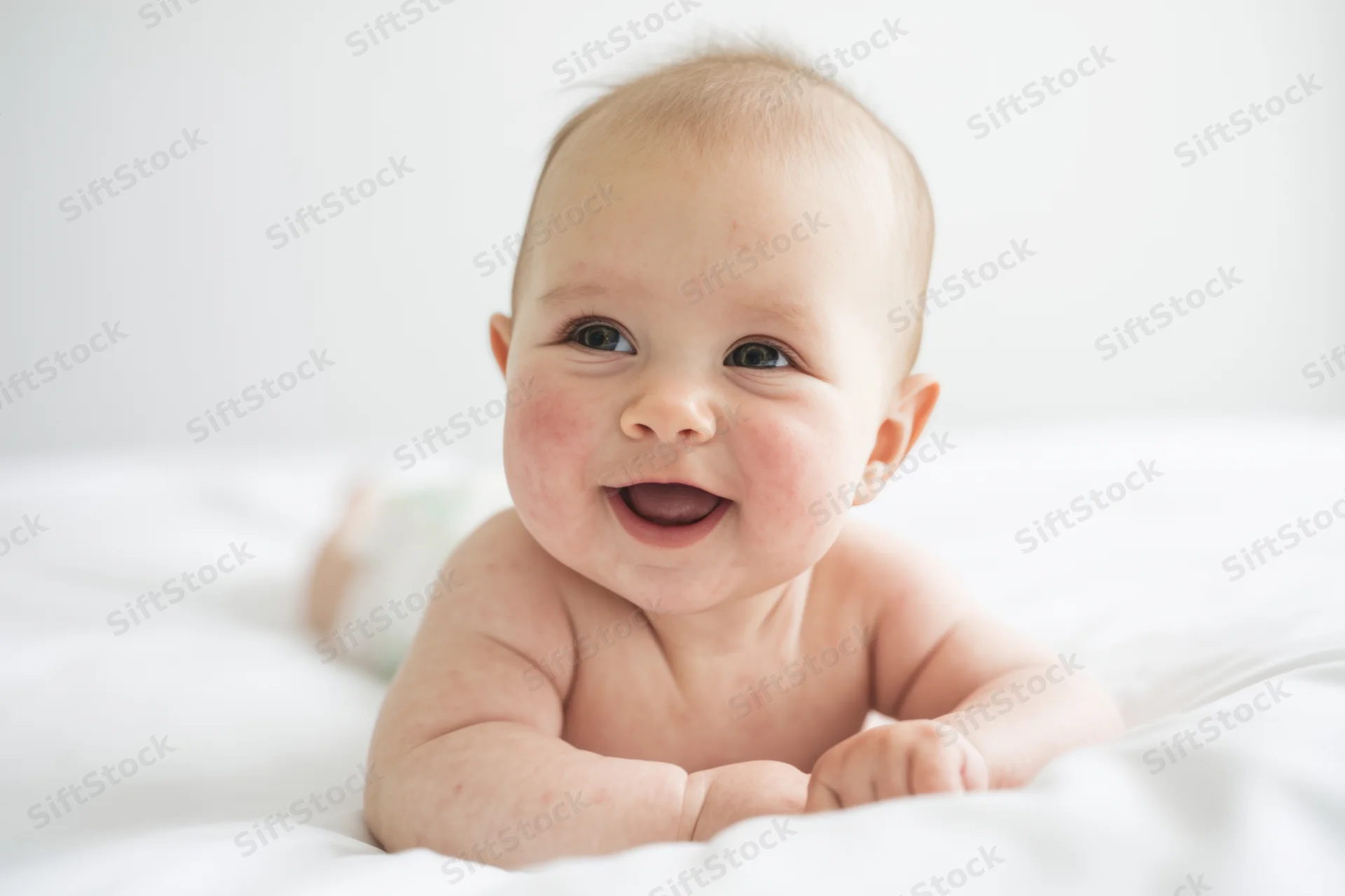 Happy baby lying on stomach on white bed smiling with rosy cheeks