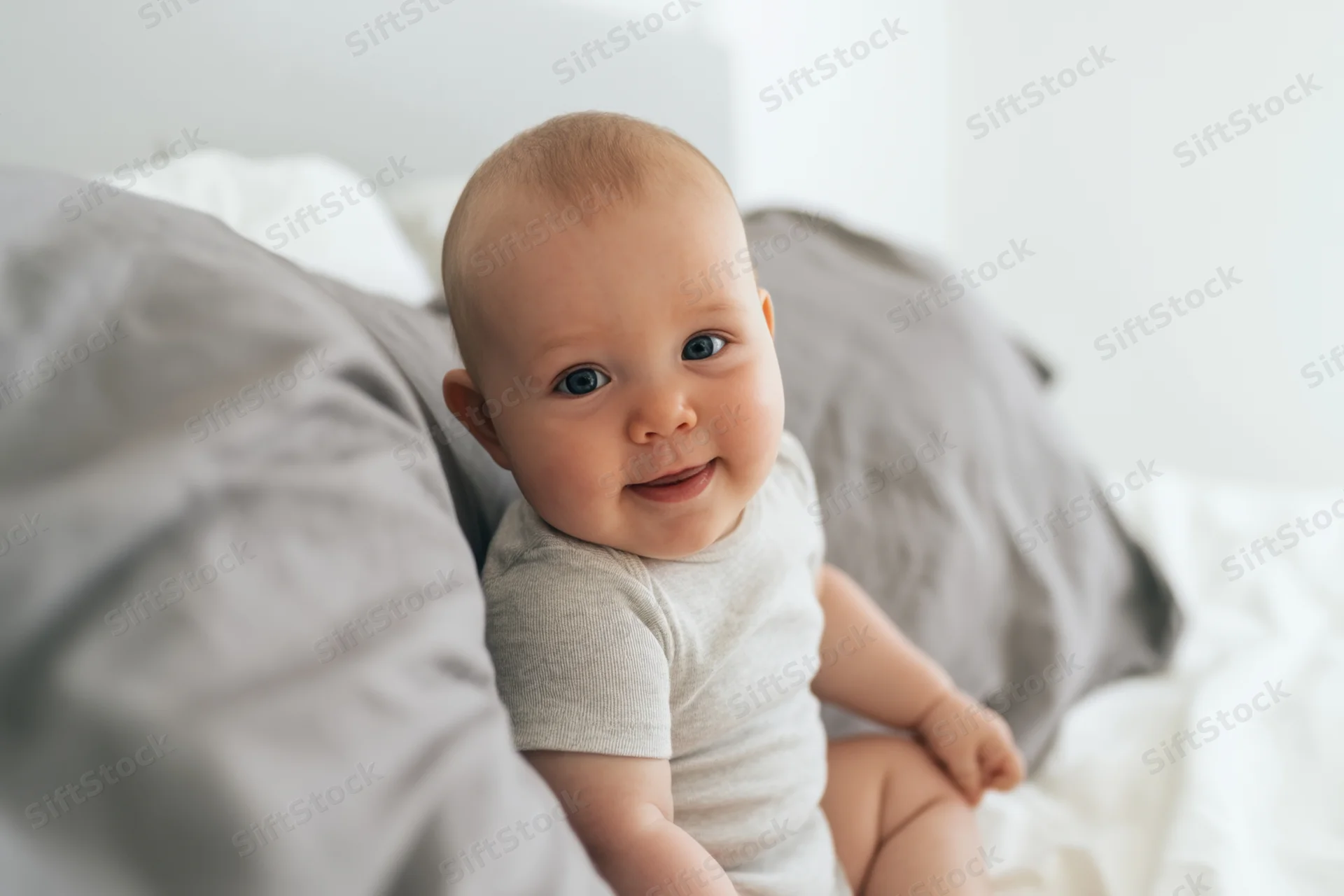Happy baby sitting on white bed with soft gray pillows wearing light gray onesie