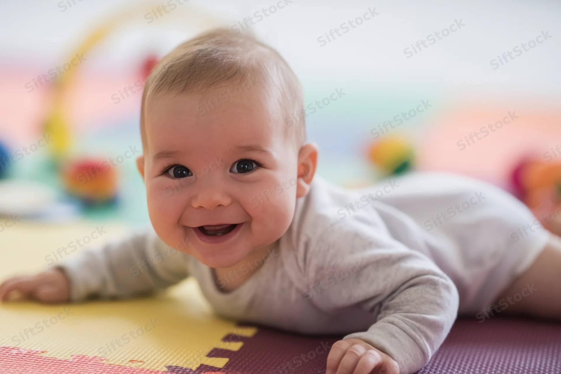 Baby lying on stomach on colorful foam mat smiling with hands on floor