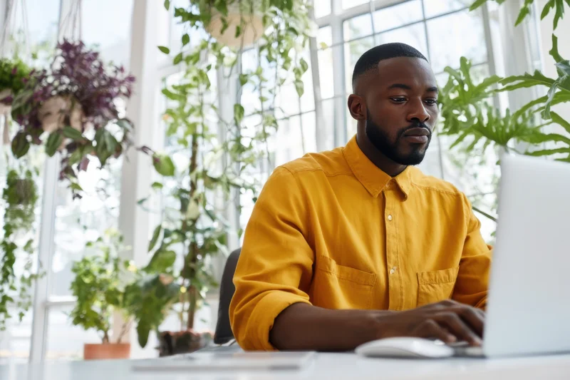 Man working on laptop at desk surrounded by indoor plants in bright office space
