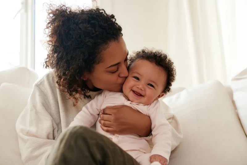 Mother kissing her smiling baby on the sofa in a bright living room