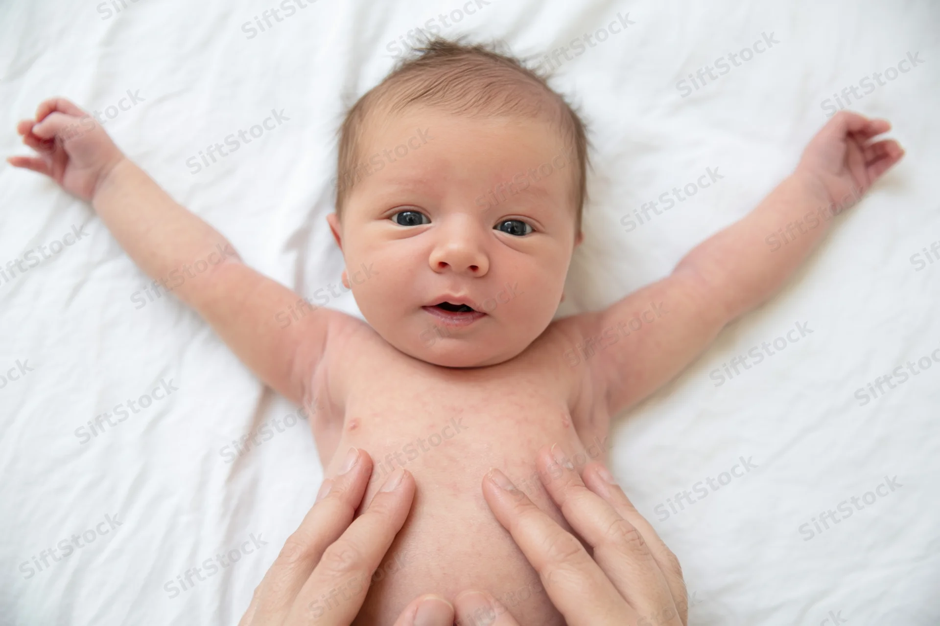 Newborn baby lying on white sheet with arms spread and hands gently touched by adult hands