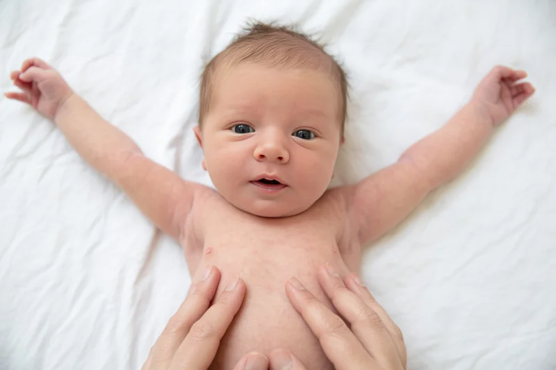 Newborn baby lying on white sheet with arms spread and hands gently touched by adult hands