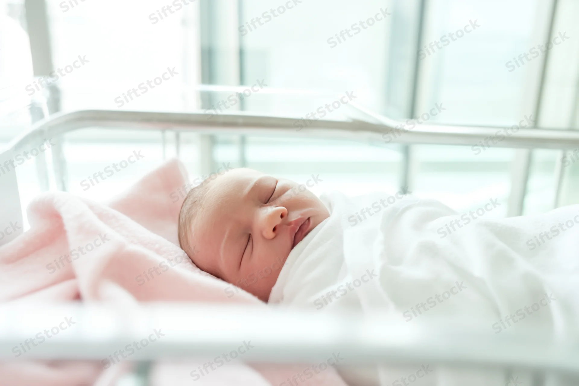 Newborn baby sleeping swaddled in white blanket in hospital bassinet with pink blanket underneath