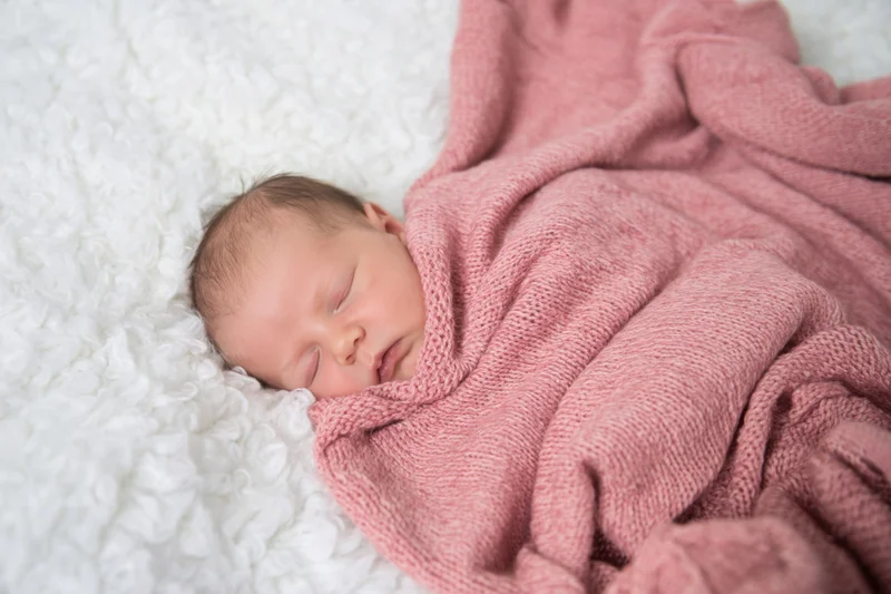 Newborn baby sleeping swaddled in pink knitted blanket on white fluffy rug