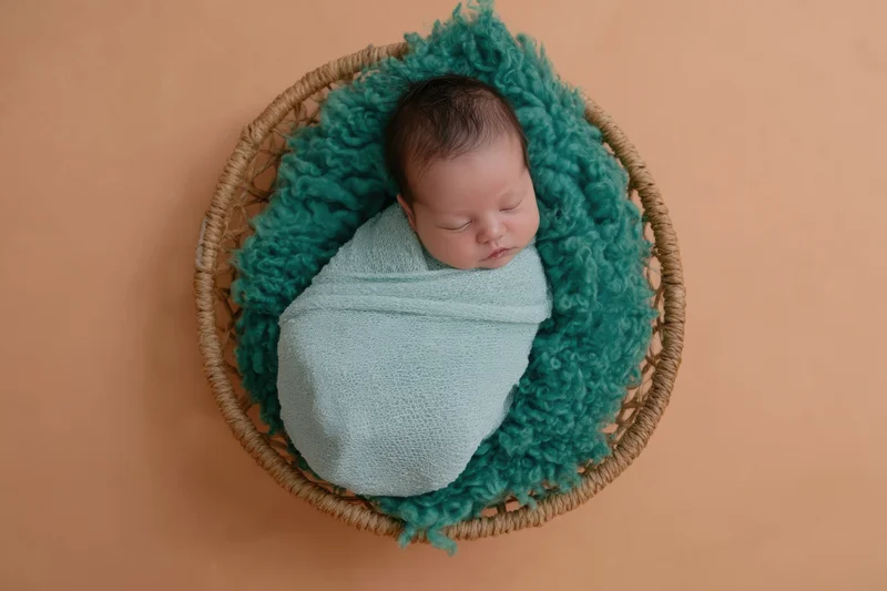 Newborn baby swaddled in pale blue wrap sleeping in wicker basket with teal fluffy blanket on peach background
