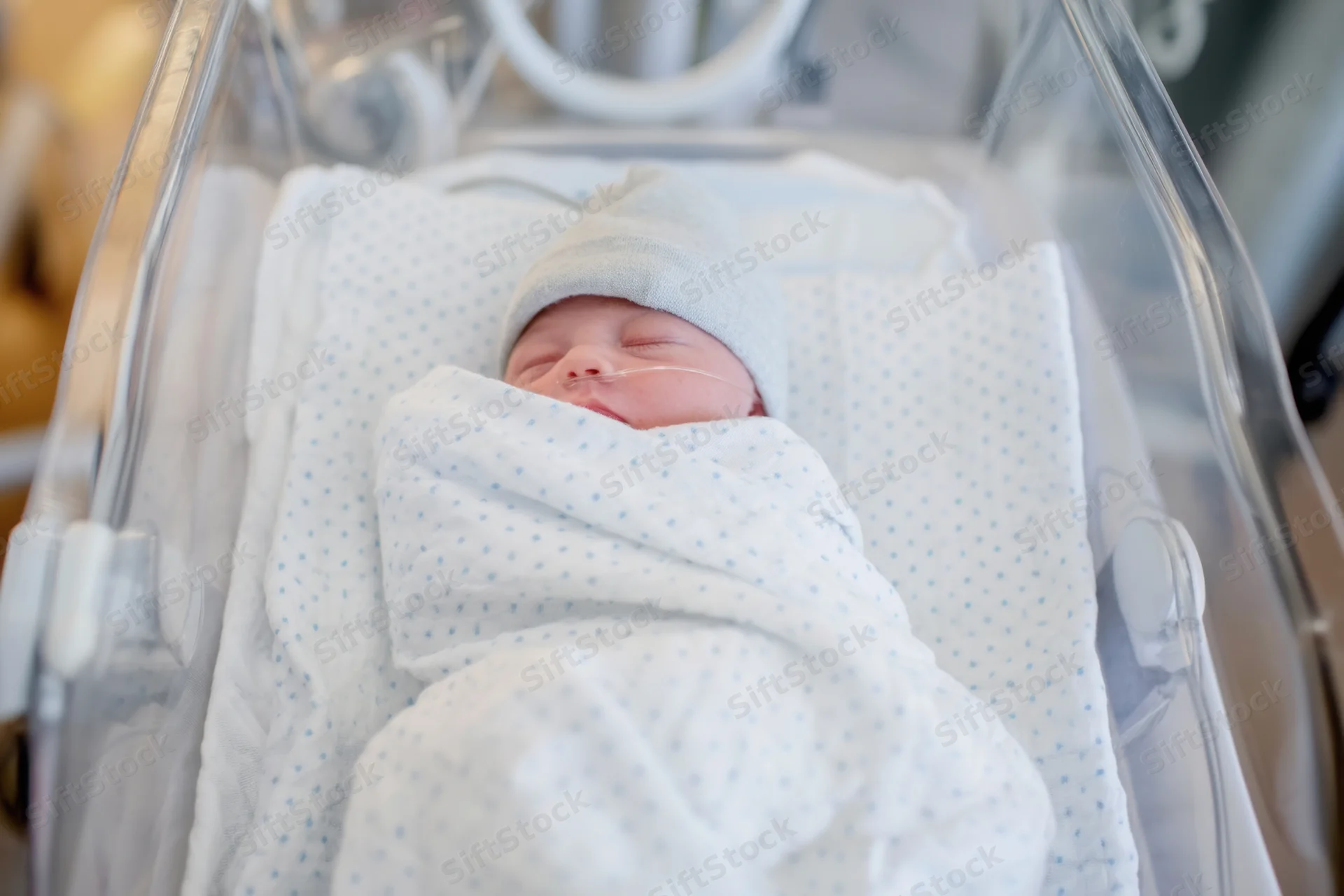 Newborn baby swaddled in white blanket with blue dots sleeping in hospital incubator