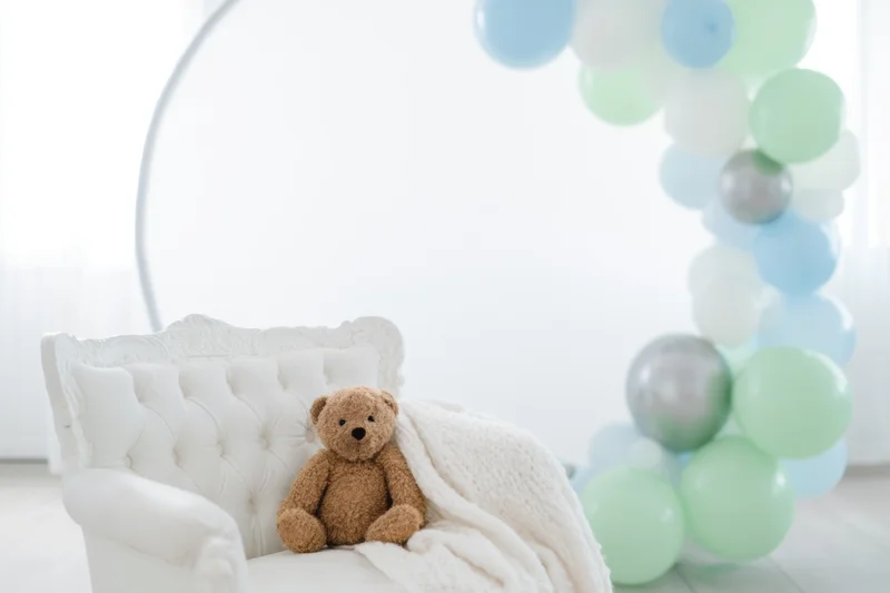 Teddy bear sitting on white tufted armchair with knitted blanket beside pastel balloon arch in bright room