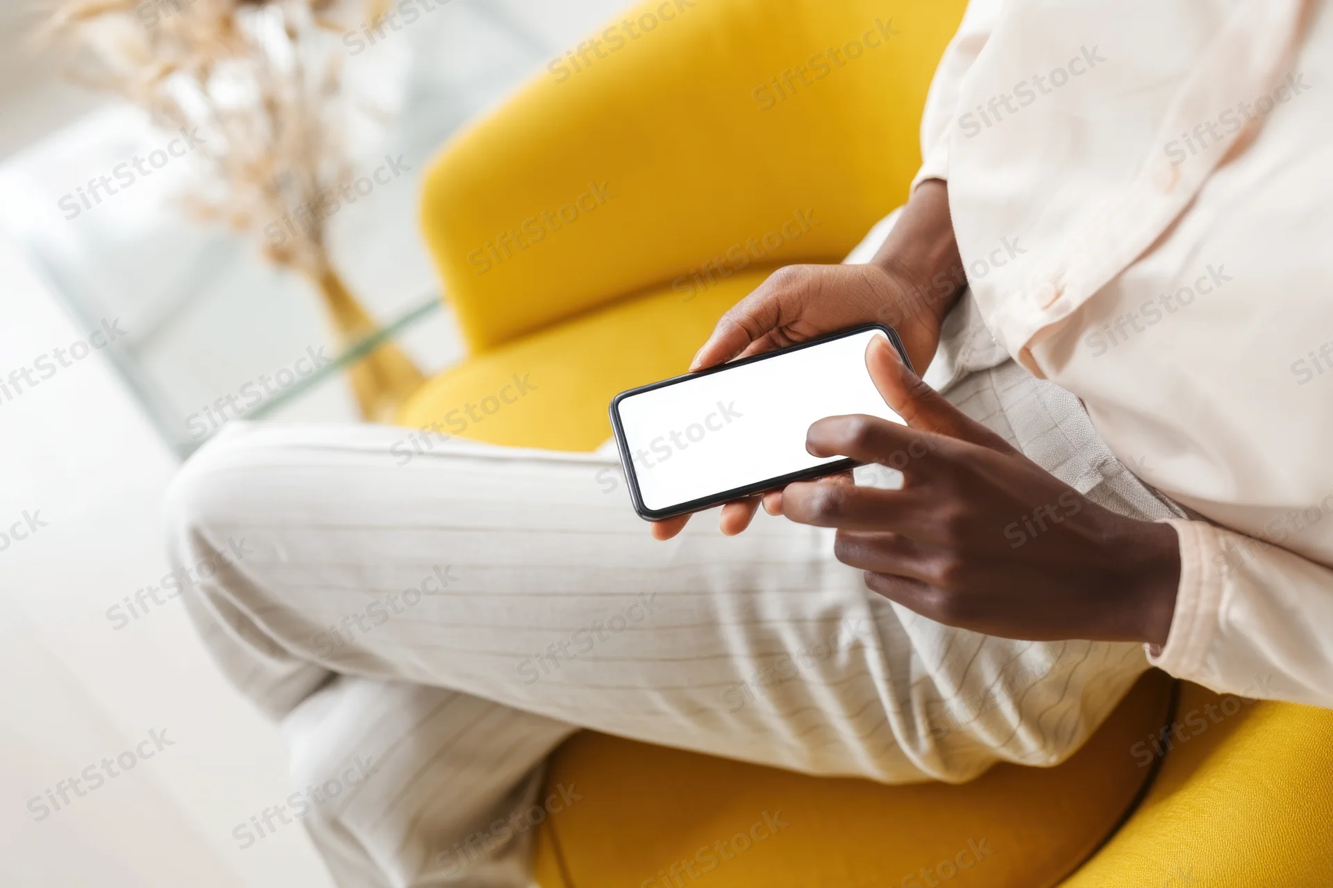 Person sitting on yellow chair using smartphone with white screen in bright modern room