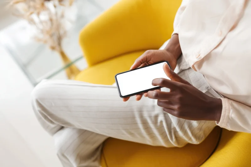 Person sitting on yellow chair using smartphone with white screen in bright modern room