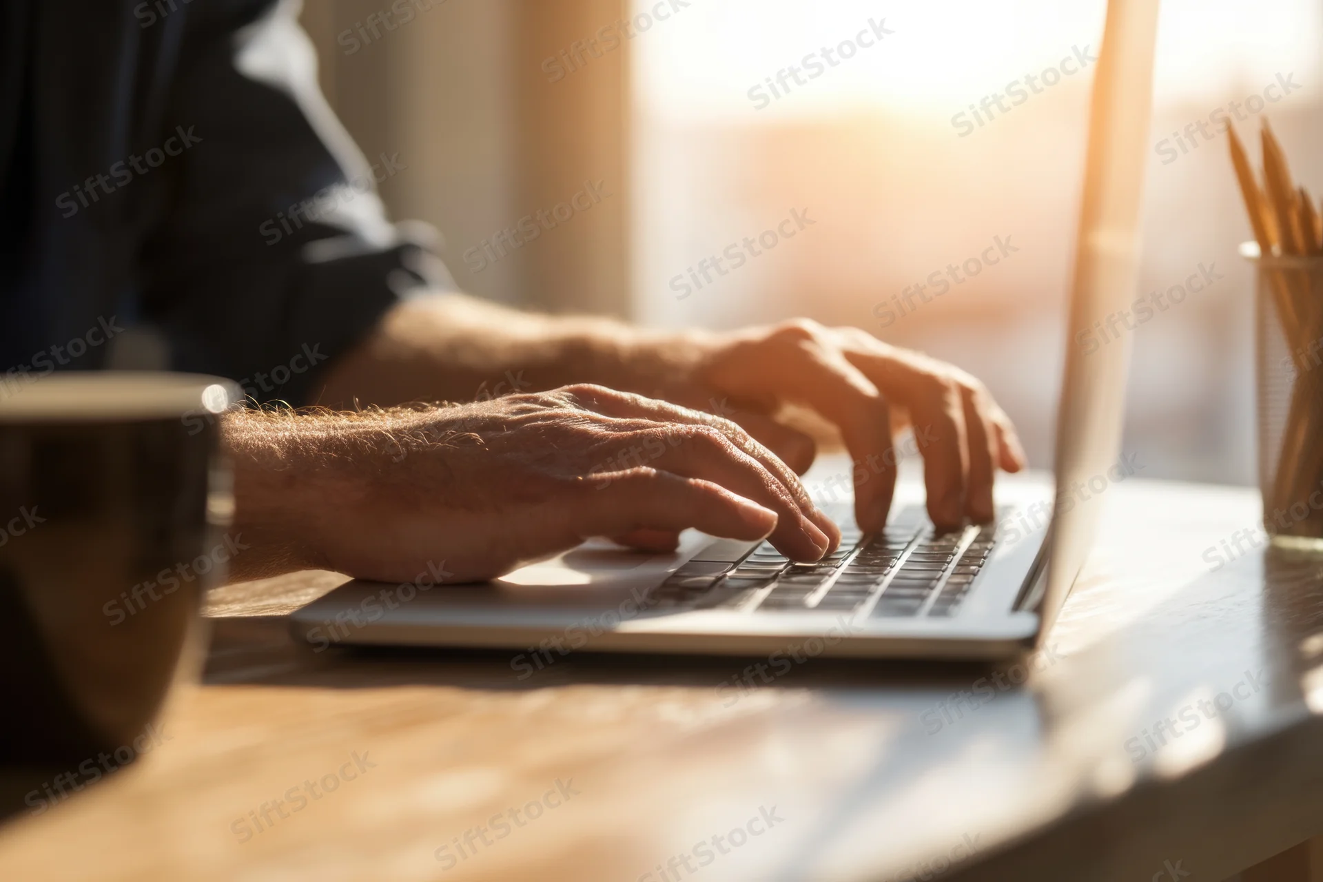 Man typing on laptop at wooden desk with coffee mug in morning sunlight