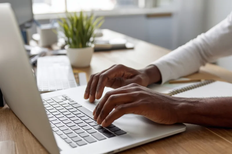 Man typing on silver laptop at wooden desk in home office setting