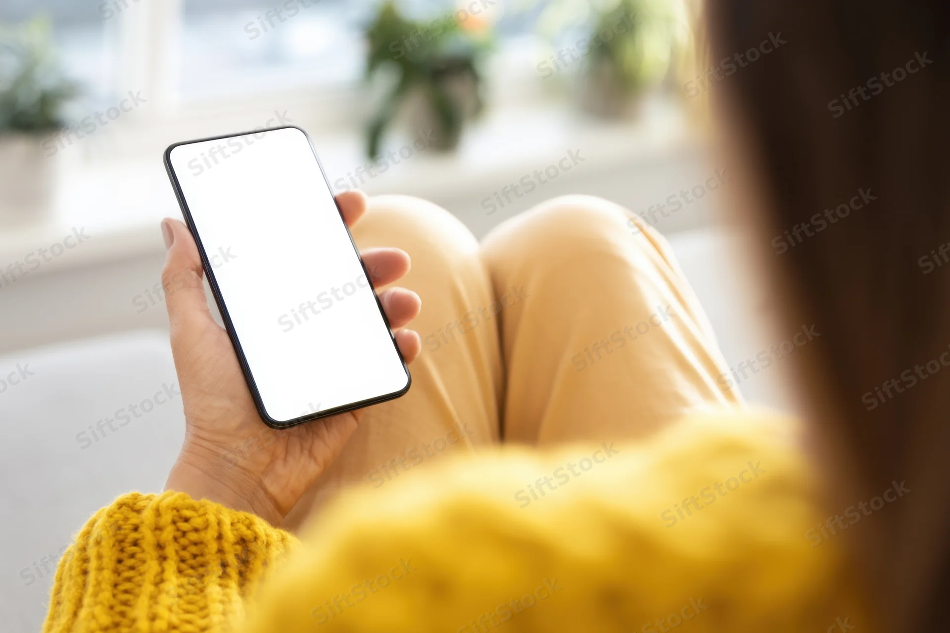 Woman holding smartphone with white screen while sitting on couch in living room wearing yellow knit sweater