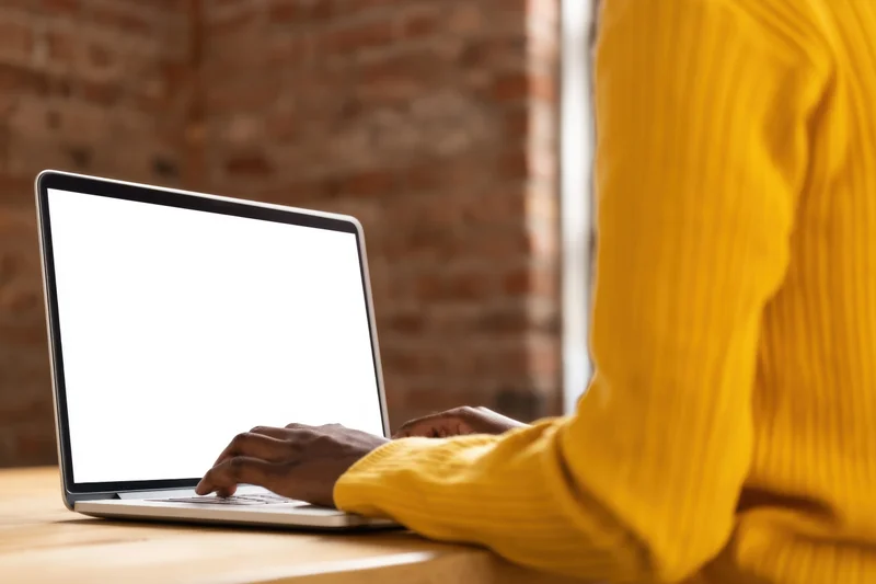 Person typing on laptop at wooden desk with yellow sweater sleeve visible