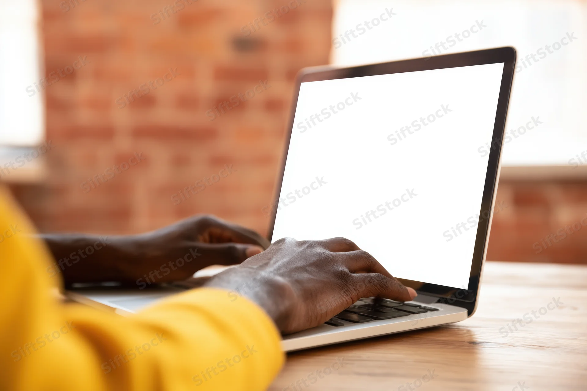 Person with yellow sleeve typing on laptop at wooden desk in brick-walled office