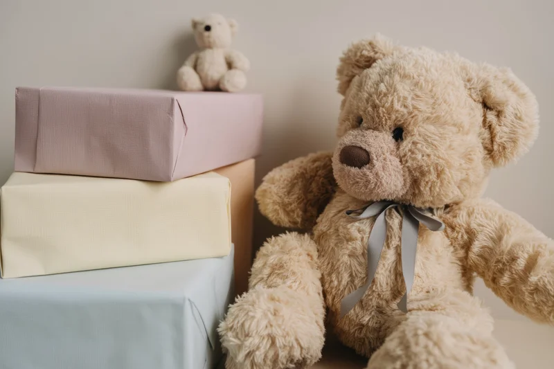 Plush beige teddy bear with grey ribbon beside stacked gift boxes on shelf
