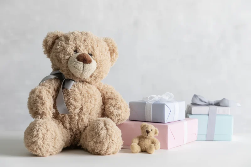 Plush beige teddy bear with gray ribbon sitting beside pastel gift boxes on white surface