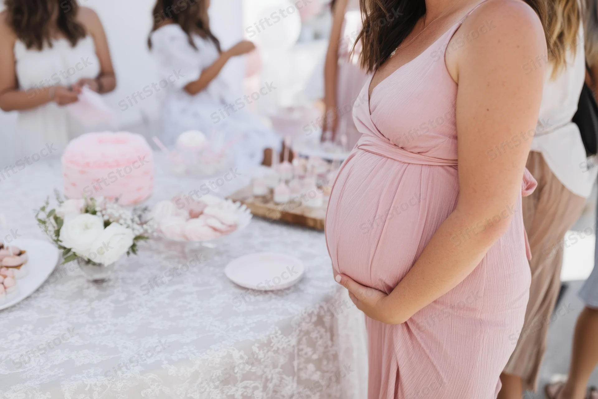 Pregnant woman in pink dress holding her belly beside a lace-covered table with desserts and flowers at a baby shower