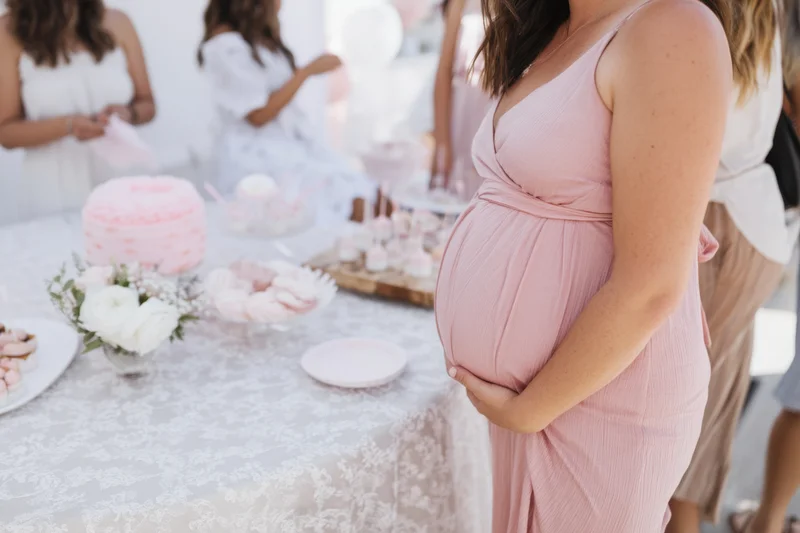 Pregnant woman in pink dress holding her belly beside a lace-covered table with desserts and flowers at a baby shower