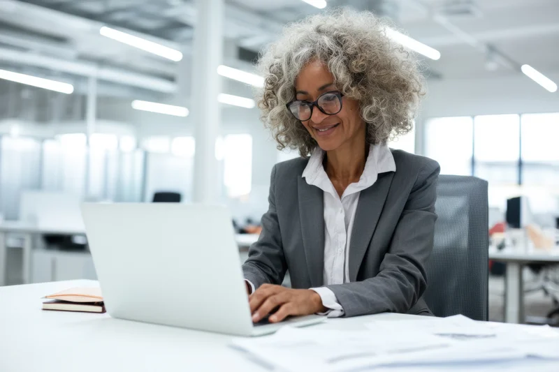 Woman in suit smiling while typing on laptop at office desk