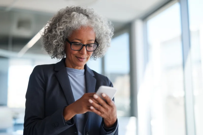 Professional woman with gray curly hair and glasses smiling while using smartphone in office