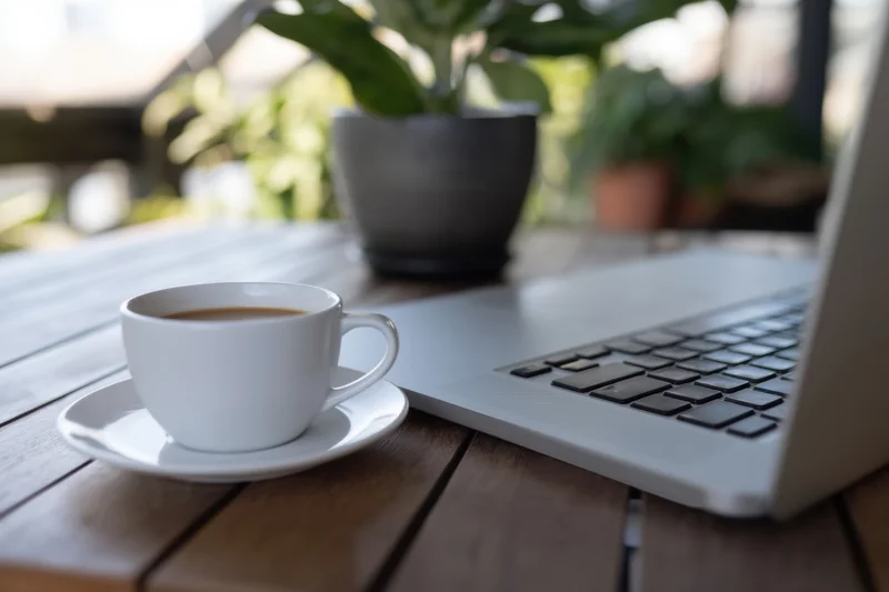 Silver laptop and coffee cup on wooden table outdoors with potted plant in background