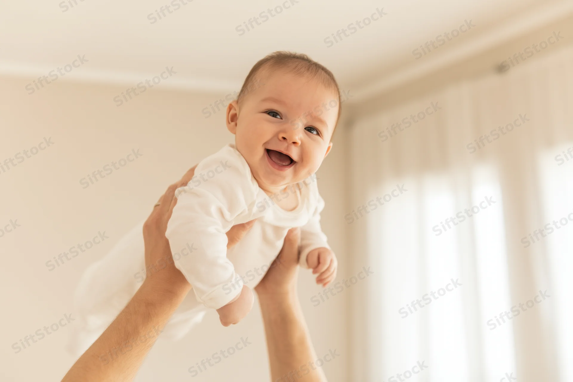 Smiling baby lifted in the air by hands in a bright room with white curtains