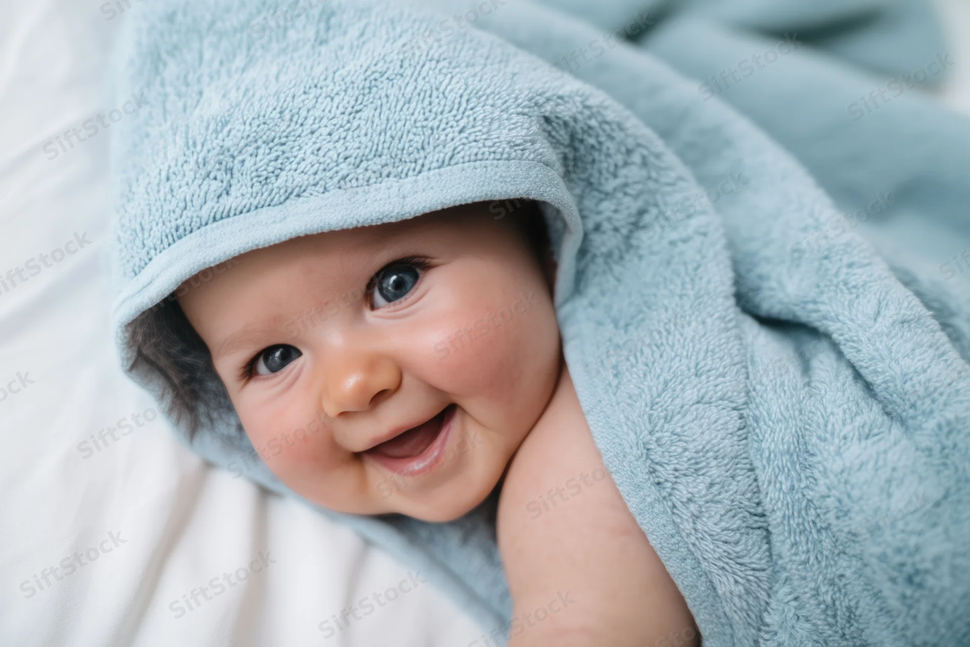 Smiling baby wrapped in blue towel lying on white bed