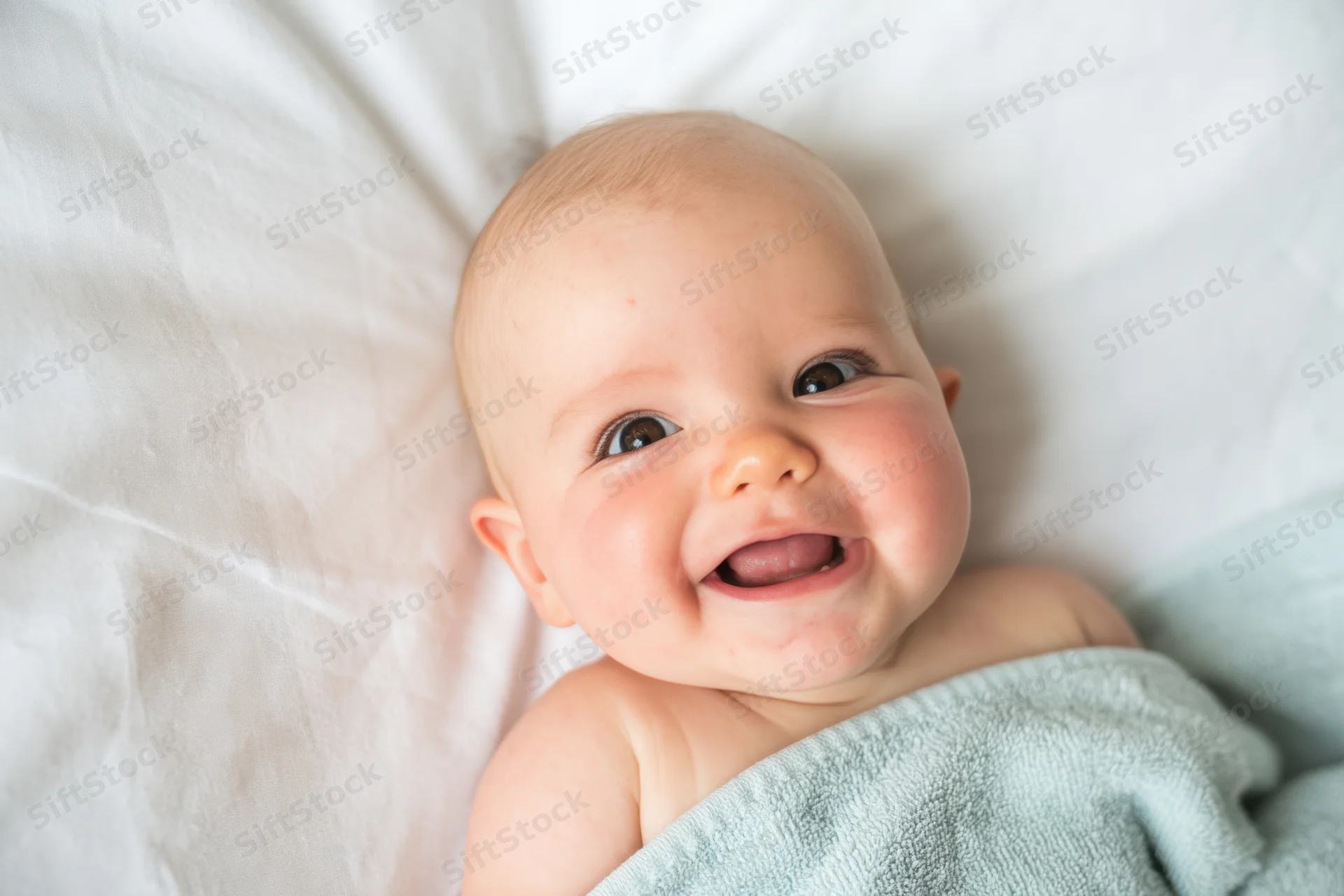 Smiling baby lying on white bed wrapped in pale blue towel