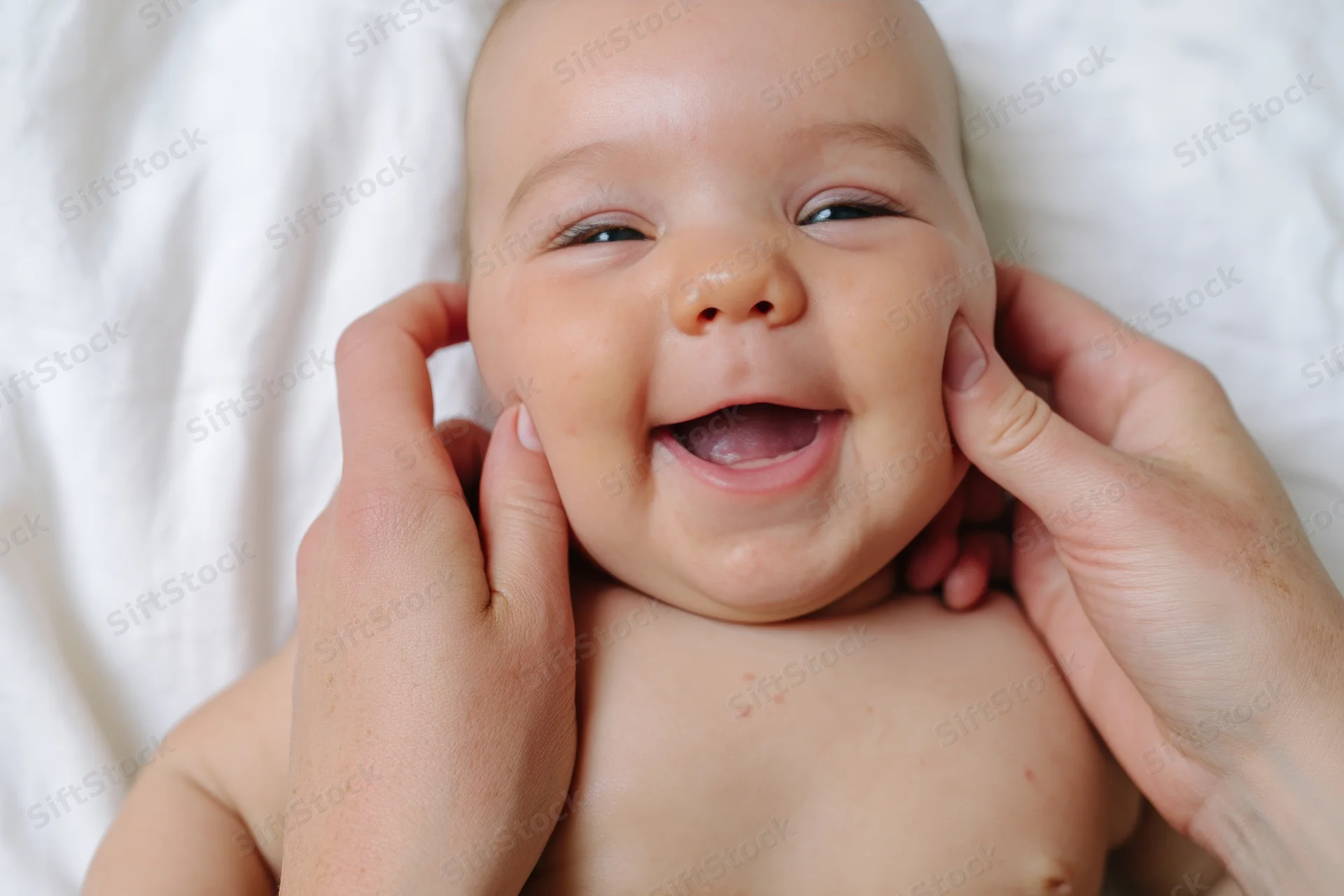 Smiling baby being gently held by hands on a soft white surface