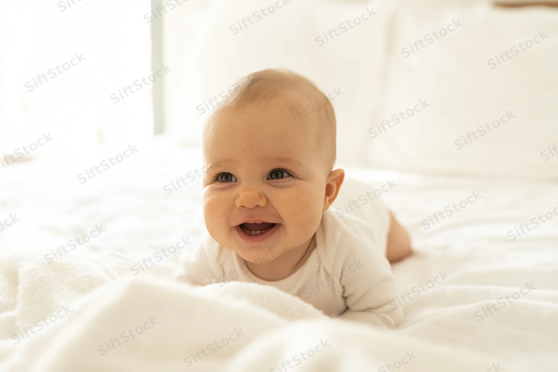 Smiling baby on tummy in white onesie on bright bed