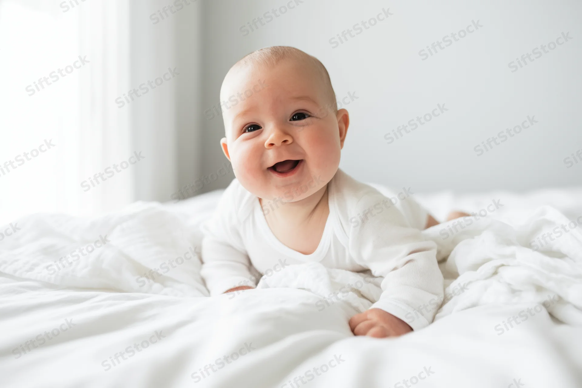 Smiling baby on tummy in white onesie on bright bed with soft blanket