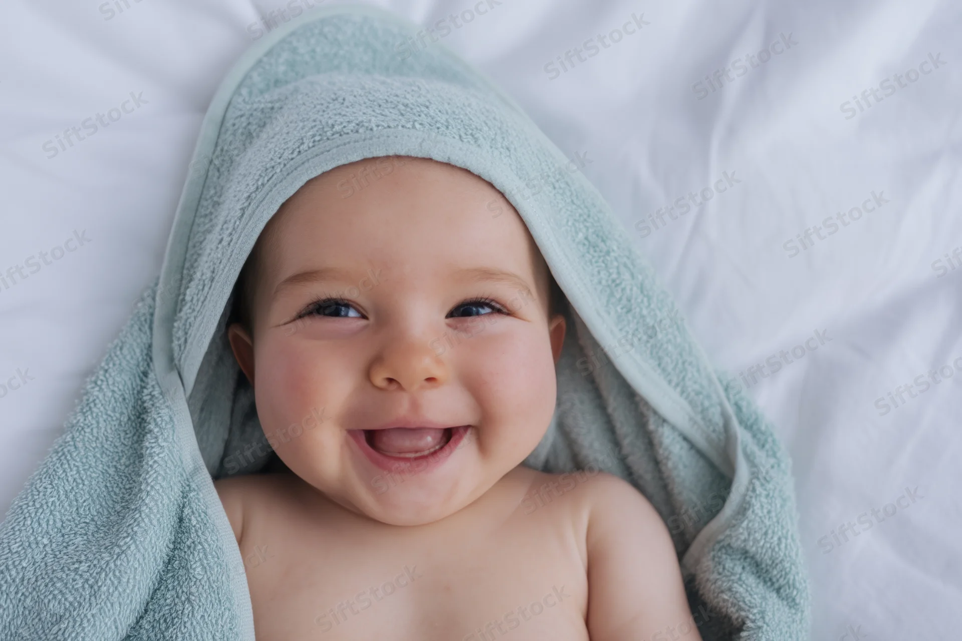 Smiling baby with pale blue towel draped over head lying on white bedding