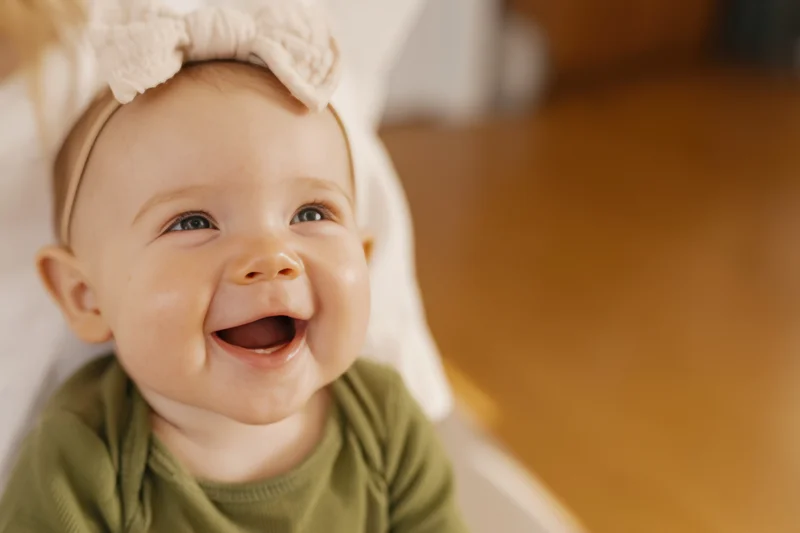 Smiling baby wearing olive green shirt and beige bow headband indoors