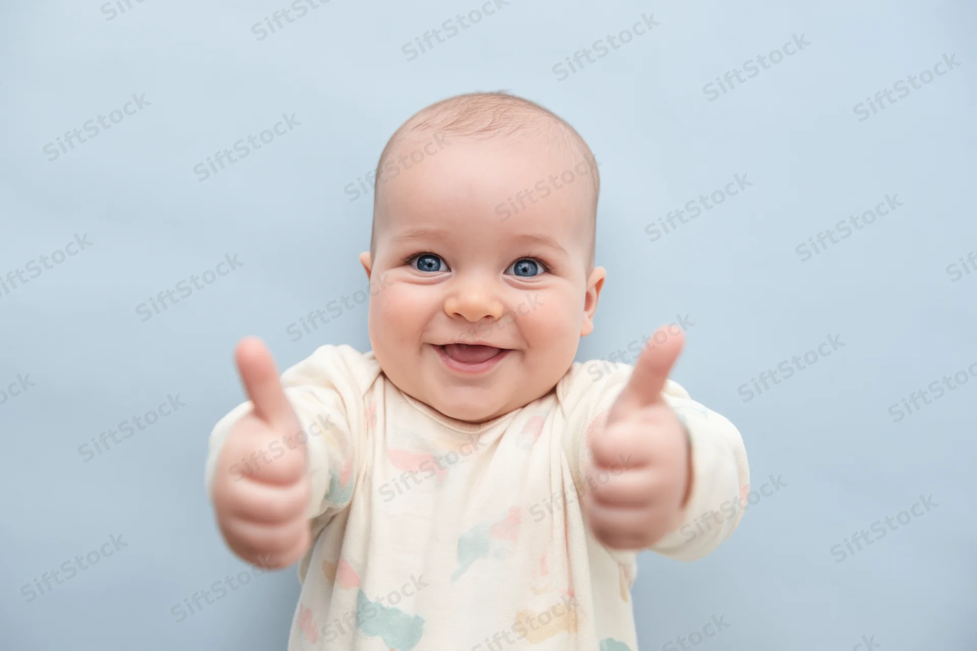 Smiling baby with blue eyes giving two thumbs up in a studio setting