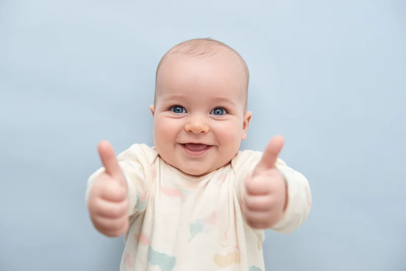 Smiling baby with blue eyes giving two thumbs up in a studio setting