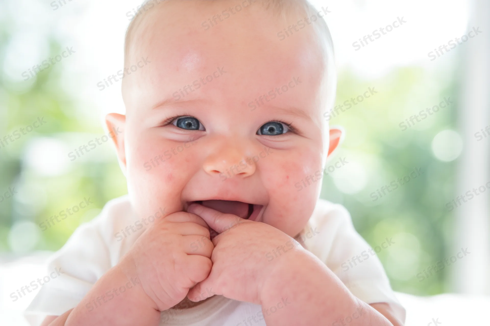 Smiling baby with blue eyes and rosy cheeks wearing white clothing, hands near mouth, close-up portrait with blurred green background