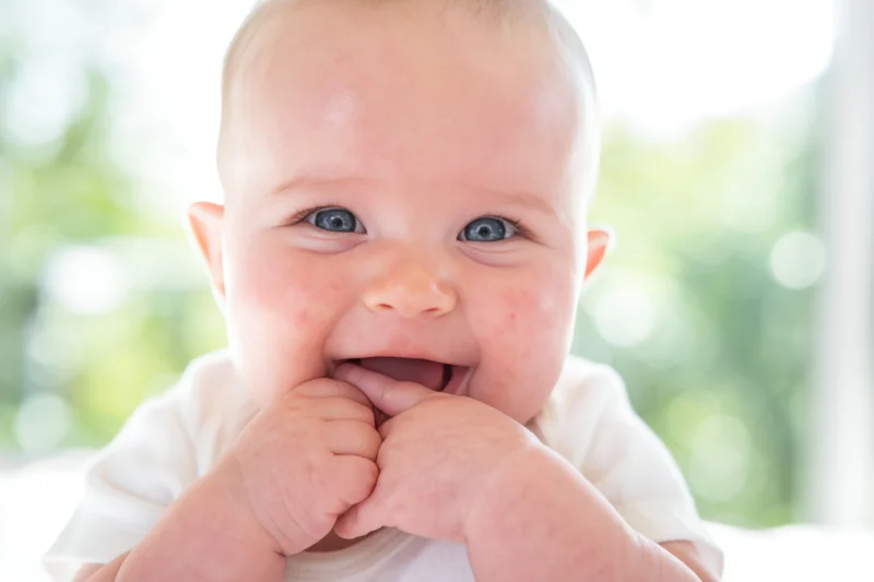 Smiling baby with blue eyes and rosy cheeks wearing white clothing, hands near mouth, close-up portrait with blurred green background