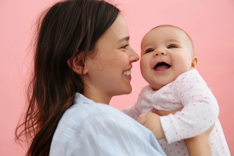 Mother smiling and cradling laughing baby against pink background