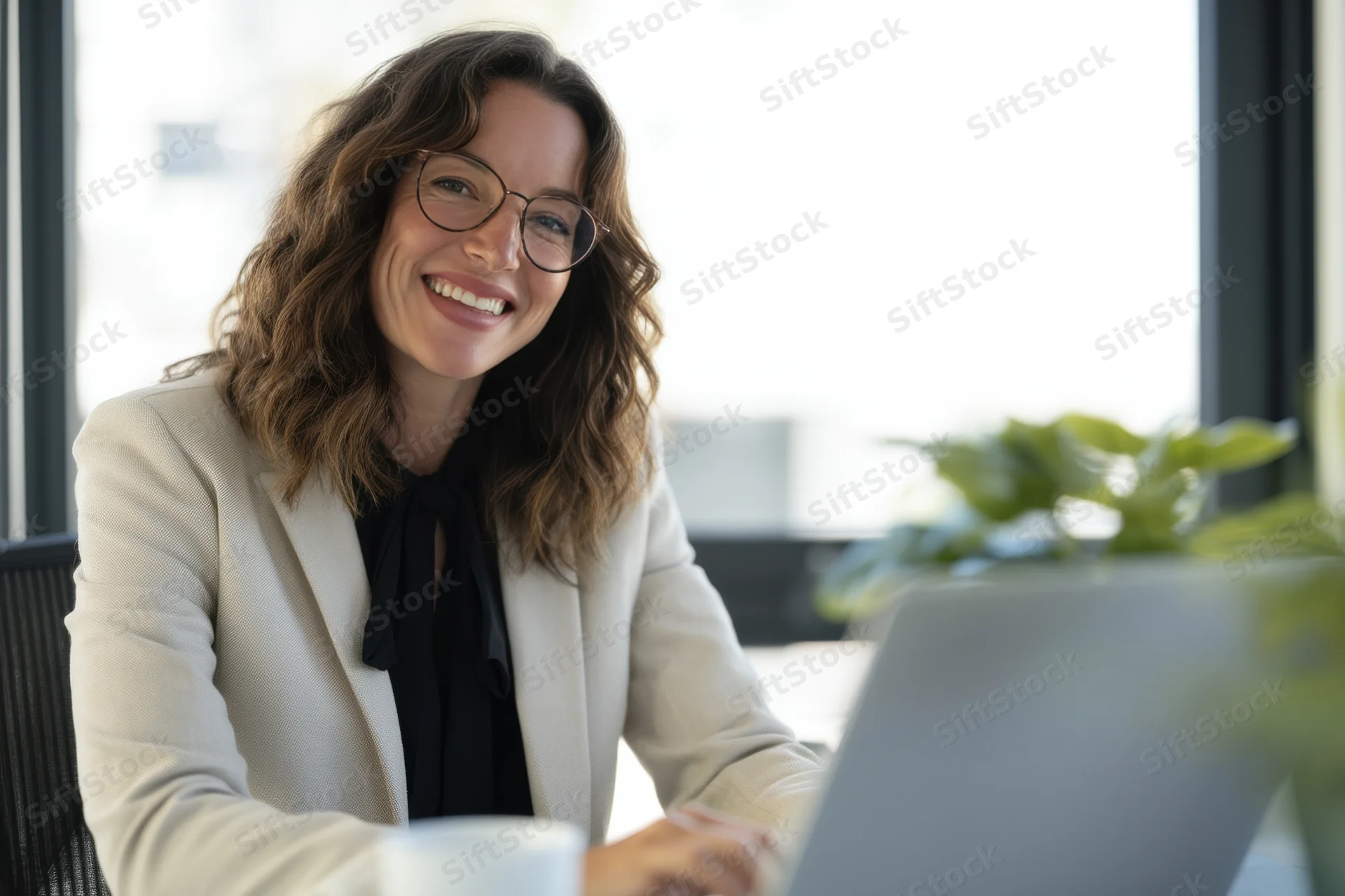 Smiling professional woman in office wearing glasses and blazer working on laptop at desk with plant nearby