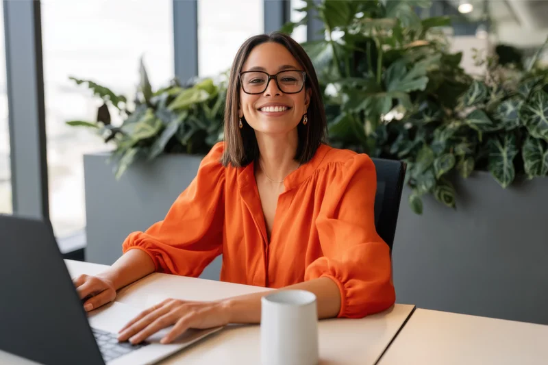 Smiling woman in orange blouse working on laptop at office desk with plants behind her