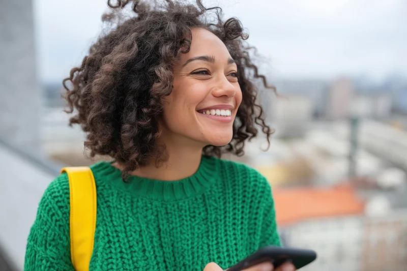 Smiling woman with natural curly hair wearing green knit sweater holding smartphone outdoors in city setting