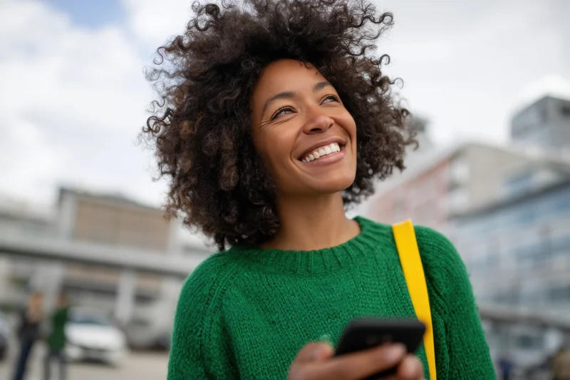 Smiling woman with natural curly hair wearing green knit sweater holding phone outdoors in city street