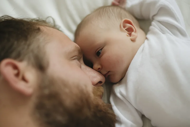 Bearded man lying close to newborn baby on bed, foreheads touching in tender moment
