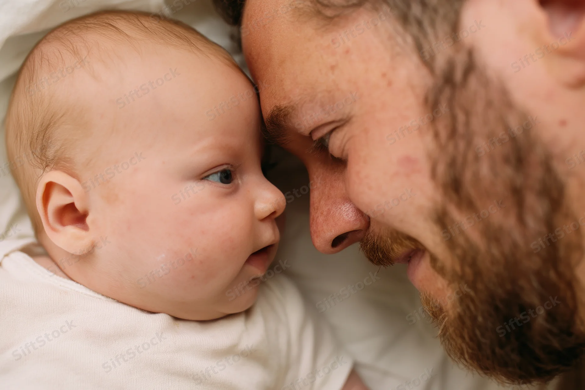 Bearded man resting forehead against baby’s head while lying close on a soft blanket