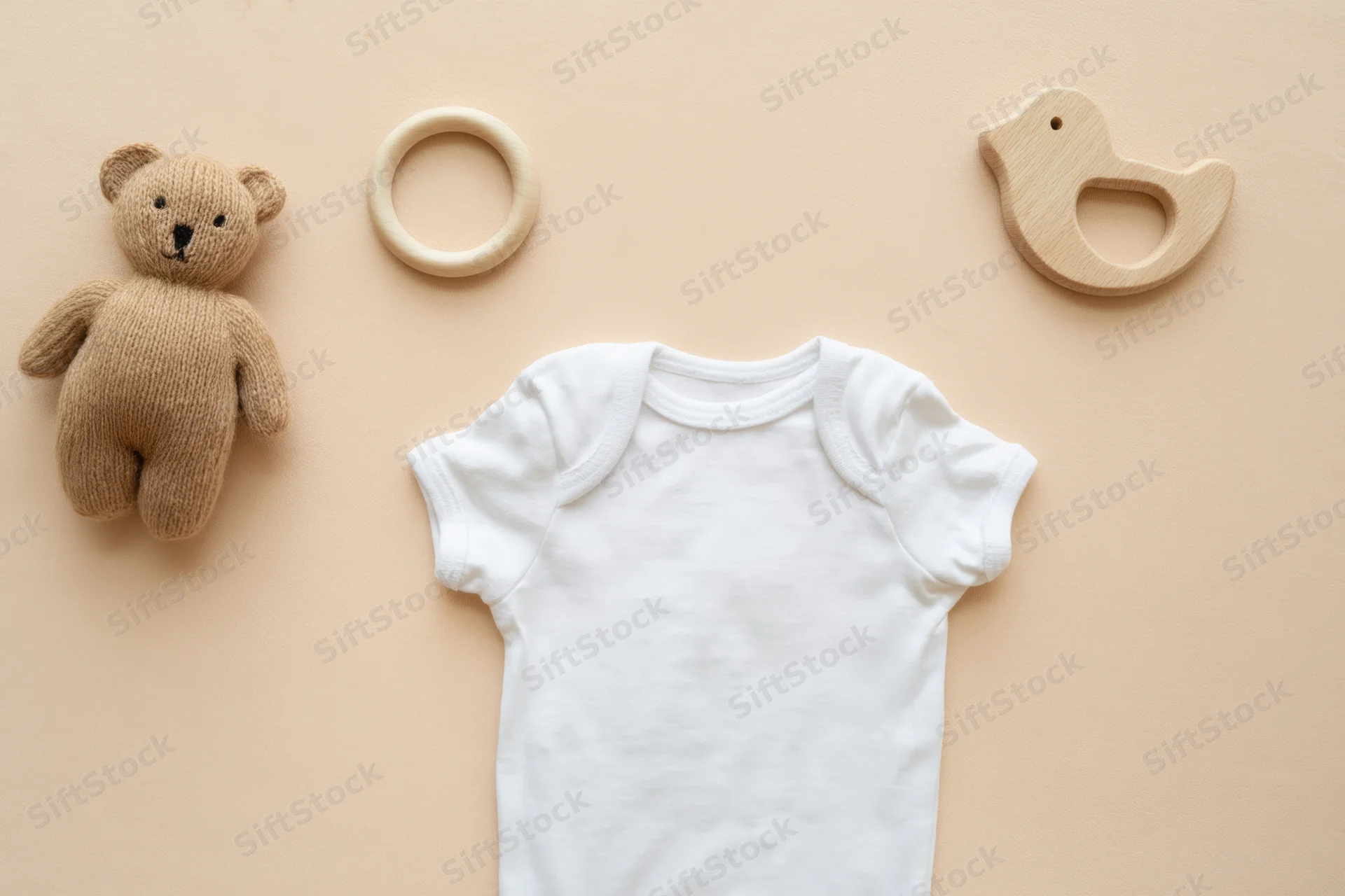 White baby onesie centered on beige background with wooden teether and plush bear toy arranged around it
